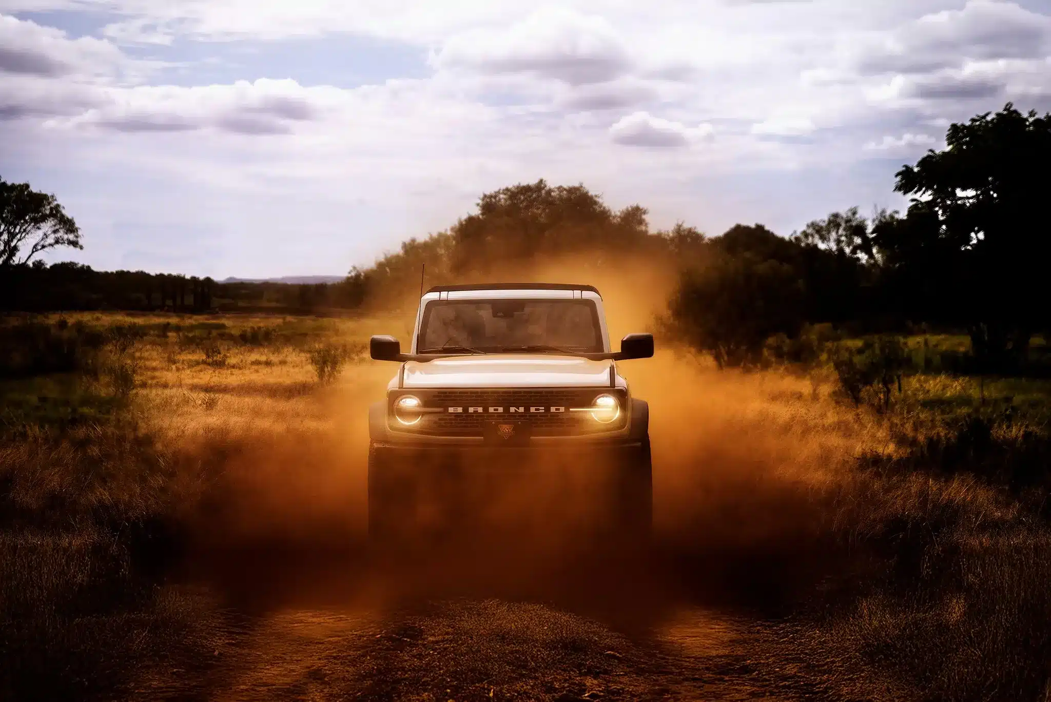 A white Ford Bronco driving through a field, kicking up a large cloud of dust.