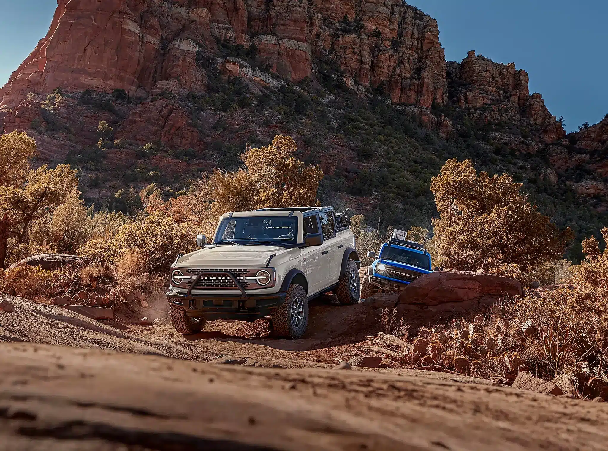 A muddy, dark-colored 2026 Ford Bronco is parked on a dirt path near a body of water and mountains. A man and a woman are walking past the vehicle carrying an orange kayak and a paddle. A green paddleboard is secured to the SUV's roof rack.