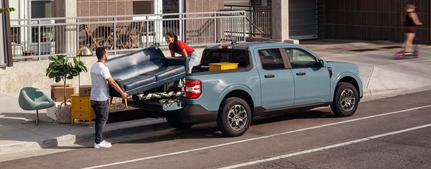 People loading a couch into the bed of a blue 2022 Ford Maverick parked by a curb.
