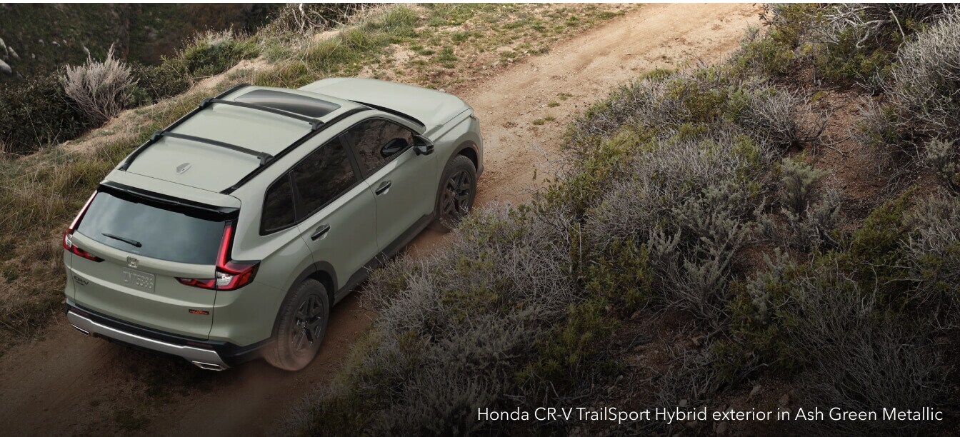 Honda CR-V TrailSport Hybrid exterior in Ash Green Metallic with roof crossbars on a dirt path.