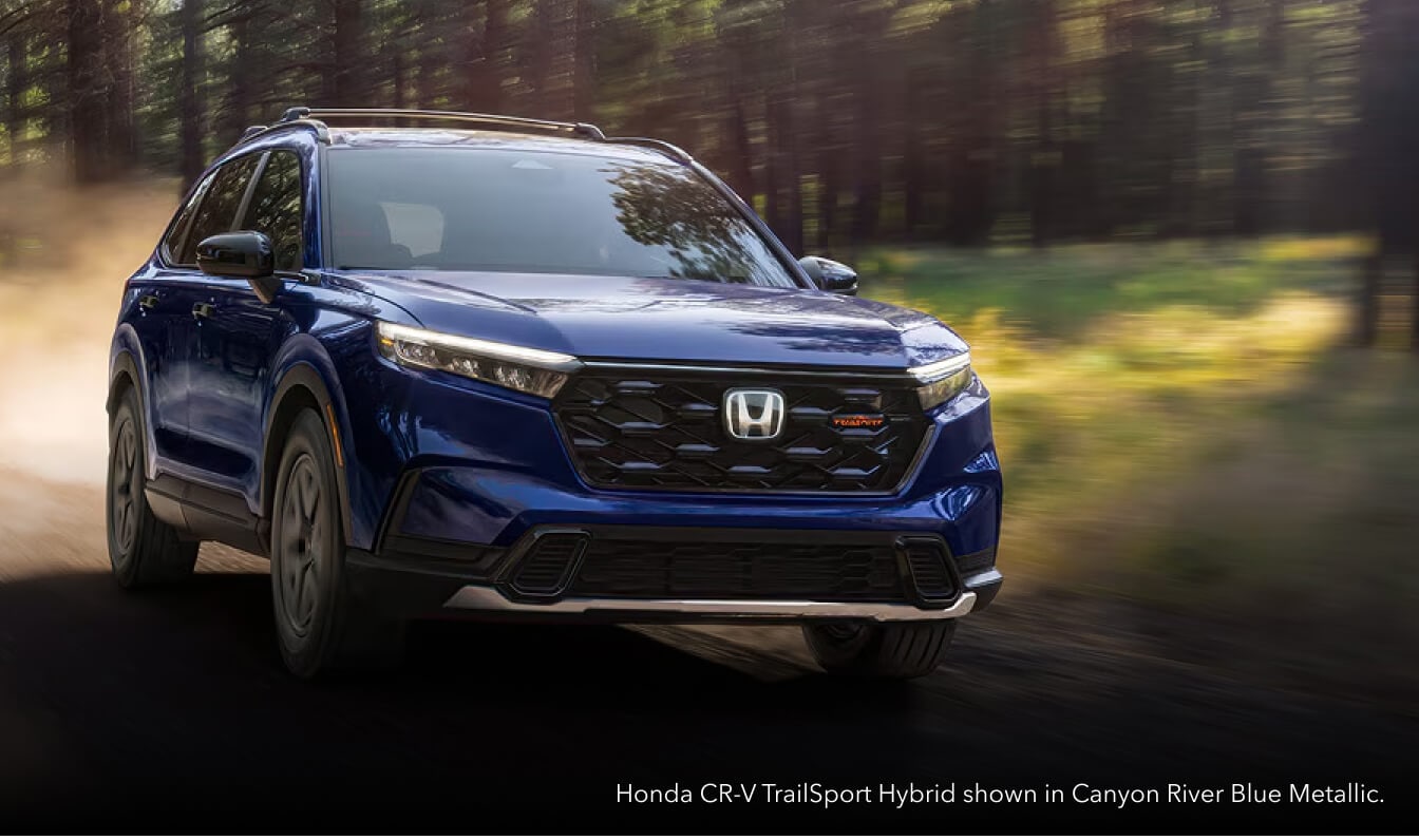 Front three-quarter dynamic motion shot of a Canyon River Blue Honda CR-V TrailSport Hybrid driving along a dusty forest trail.