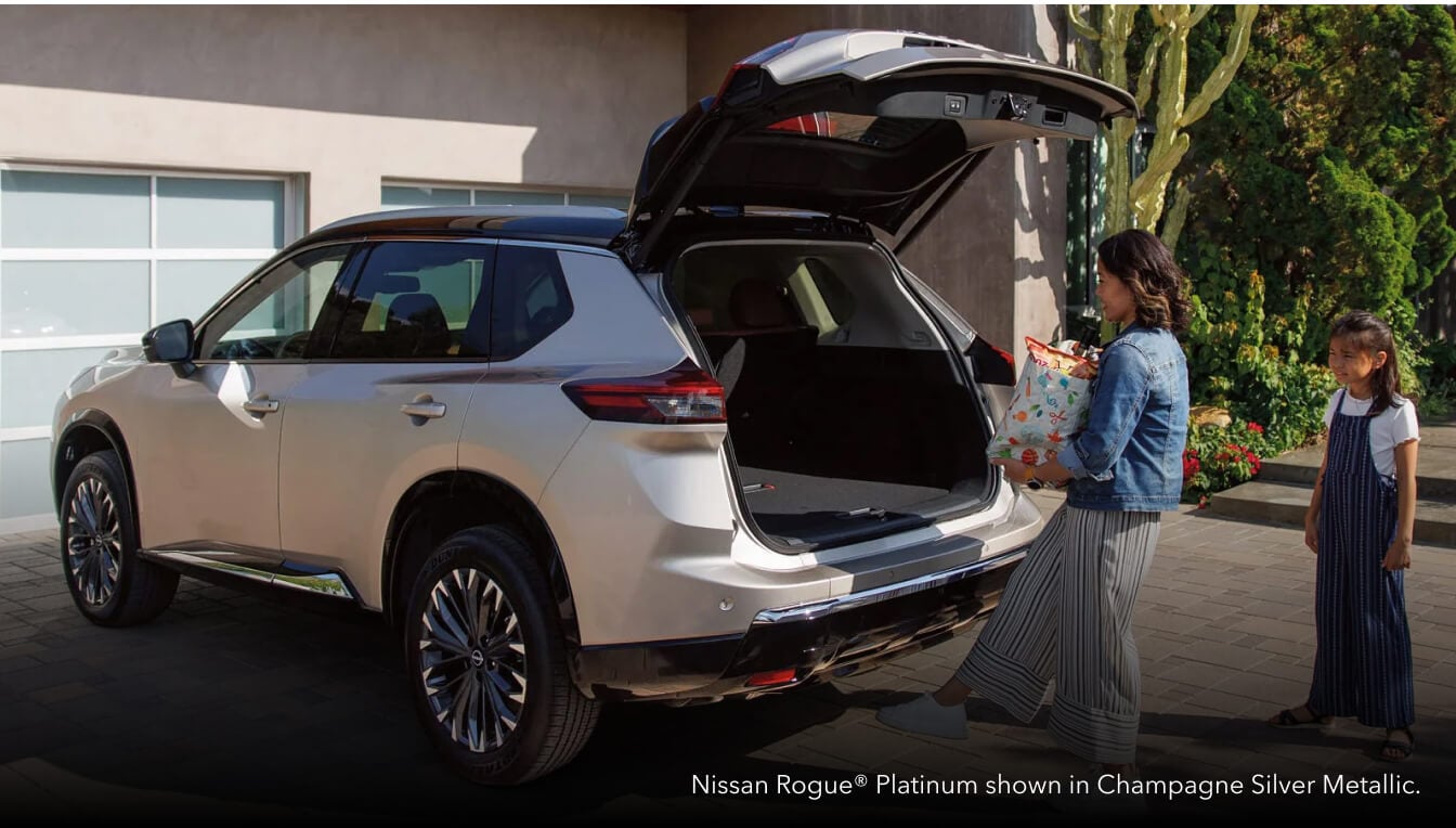 Rear three-quarter view of a Champagne Silver Nissan Rogue Platinum with an open liftgate as a woman activates the kick sensor.