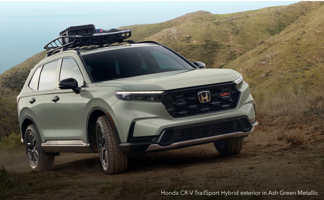 Honda CR-V TrailSport Hybrid exterior in Ash Green Metallic with roof cargo basket on a dirt trail.