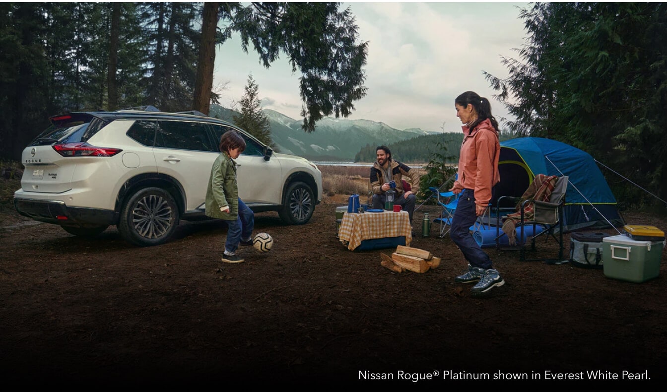Rear three-quarter view of an Everest White Nissan Rogue Platinum parked at a wooded campsite with a family.