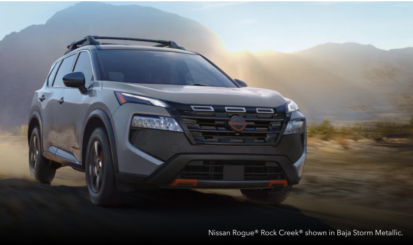 Front three-quarter dynamic motion shot of a Baja Storm Metallic Nissan Rogue Rock Creek driving along a dusty desert path.