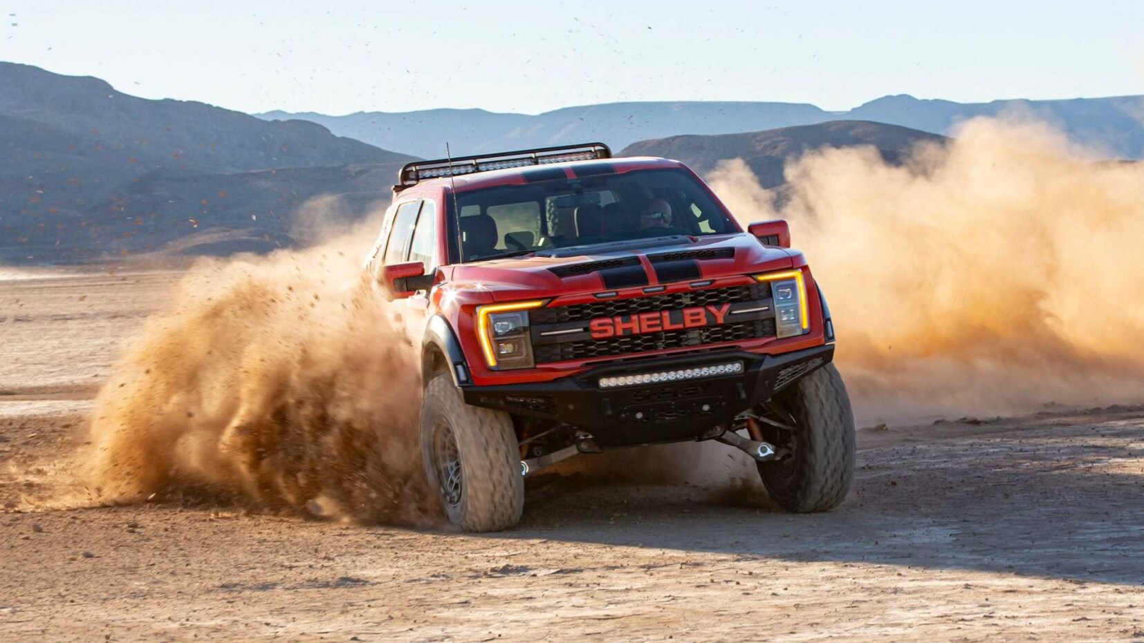 A front view of a cherry red with black racing stripes 2022 Ford Shelby Baja Raptor F-150 as it takes a hard turn kicking a cloud of dust into the air with mountains in the backgroun