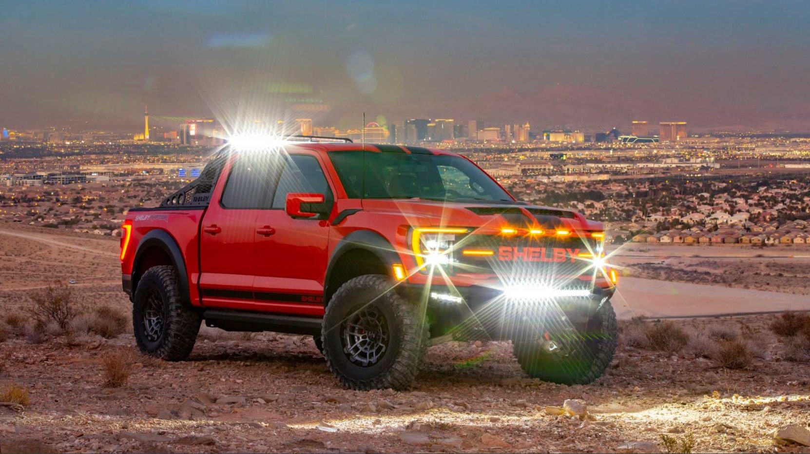 A bright red with black racing stripes 2022 Ford Shelby Baja Raptor F-150 sits parked on a hill at dusk with its lights flaring and Las Vegas in the background