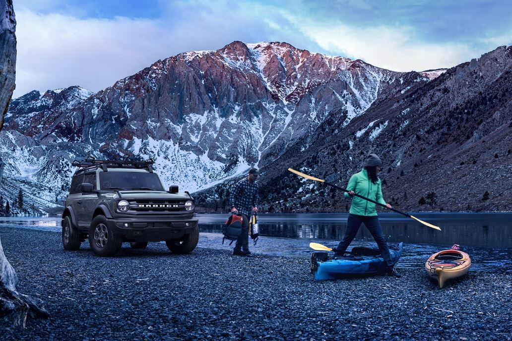 A Ford Bronco parked near a river with mountains in the background.