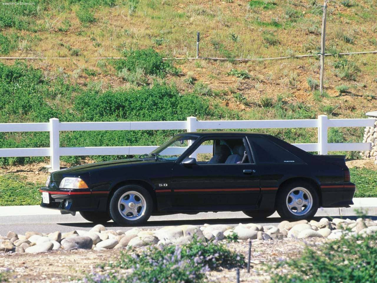 A black 1987 Ford Mustang parked in front of a white fence.