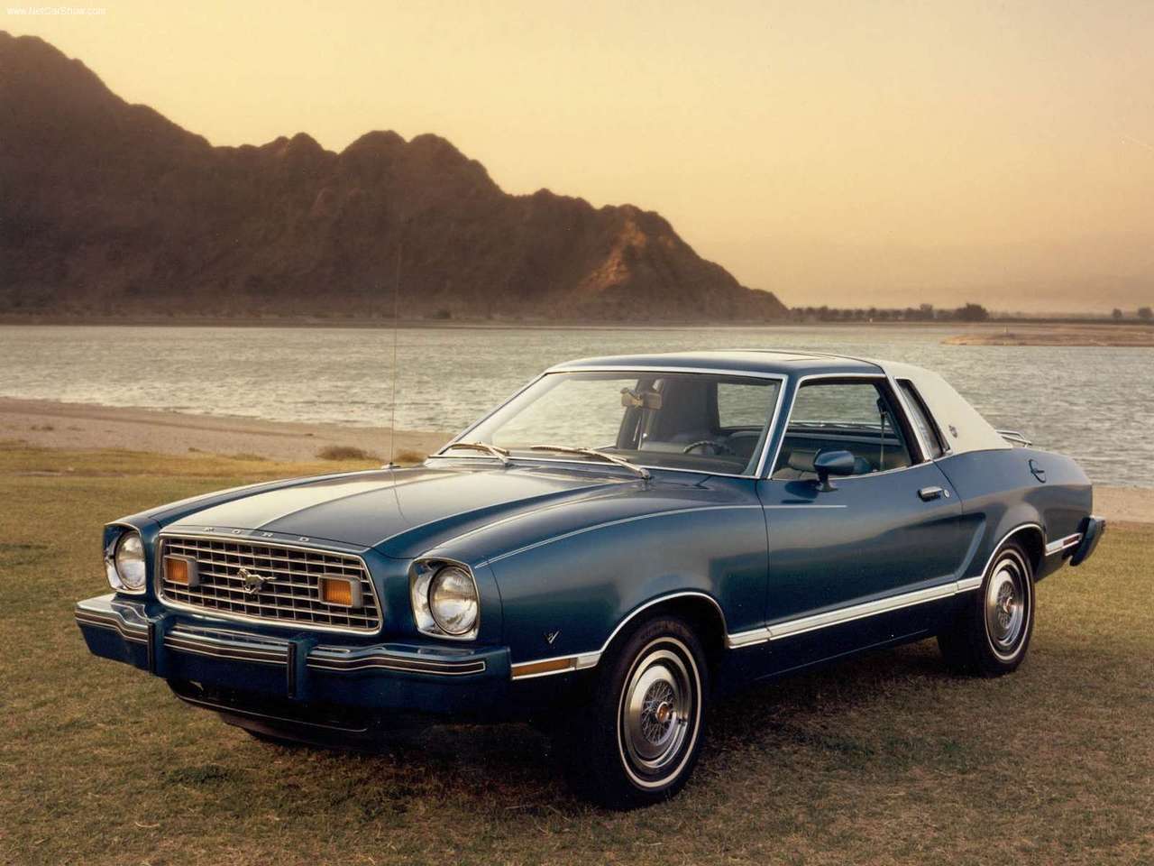 A 1977 Ford Mustang parked on a beach with mountains in the background.
