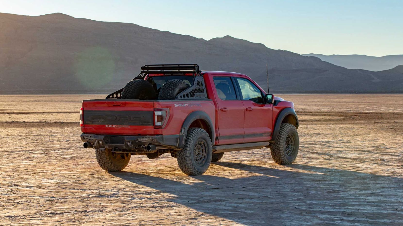 A view of the 2022 Ford Shelby Baja Raptor F-150 in cherry red with black racing stripe from the side and behind, as it is parked on an angle in the desert where the sun is setting with two spare wheels in the truckbed
