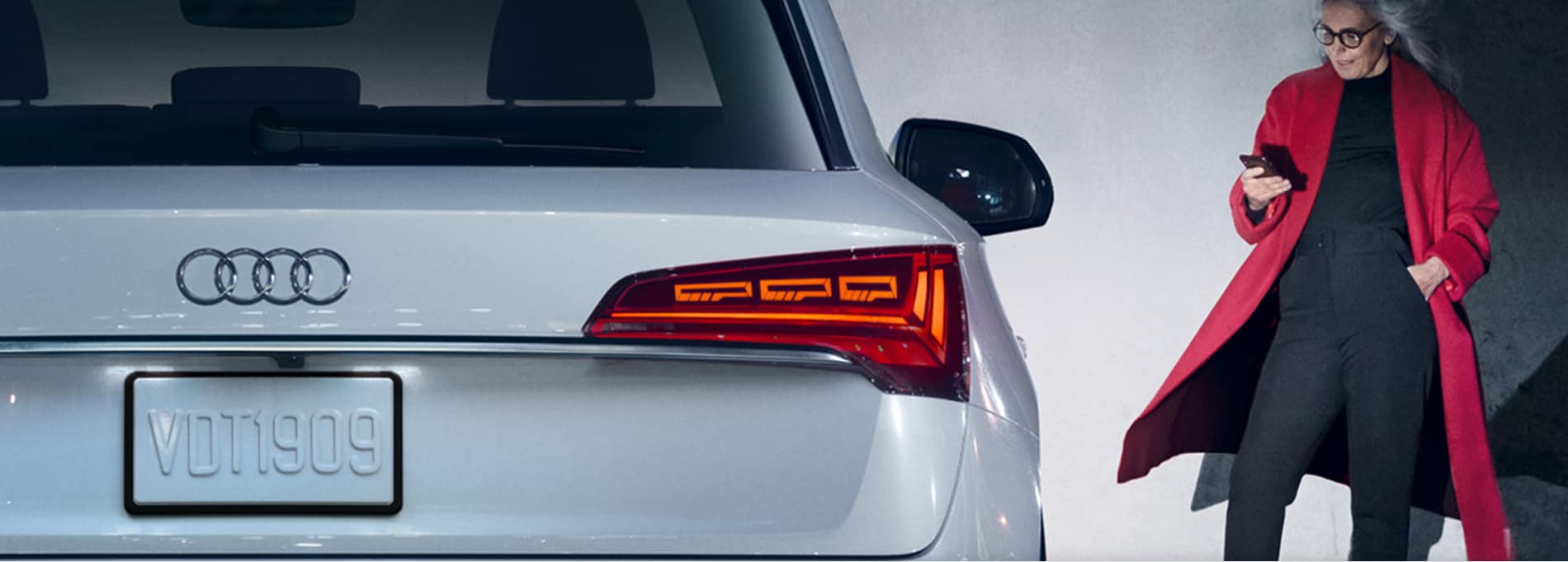 A woman in a red trench coat checks her smartphone in the background while the tail lights and right rear fender of a 2023 Audi Q5 are shown in the foreground.