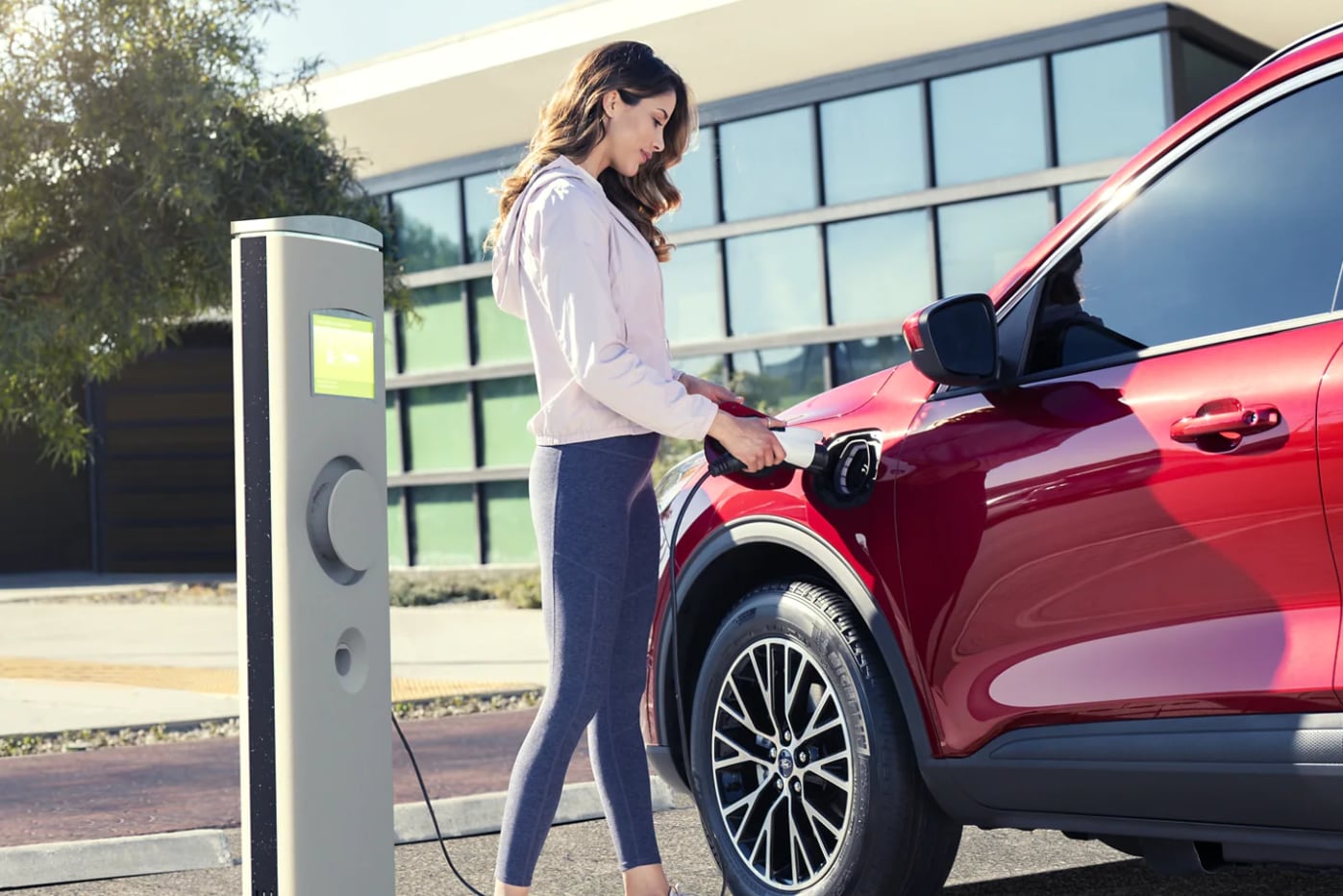 A woman in a white blouse and jeans charges her red new electric vehicle at an outdoor charging station with modern buildings in the background on a sunny day