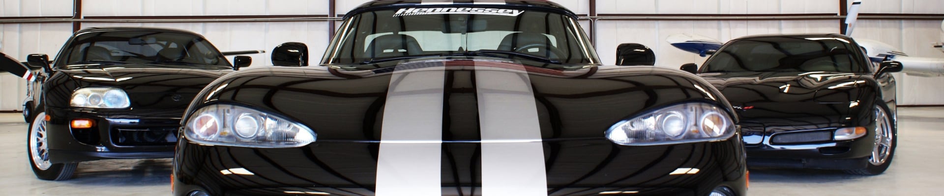 Black used Dodge Viper with 2 gray racing stripes center middle in front of a black used Corvette and a black used Acura Integra parked in an airplane hangar