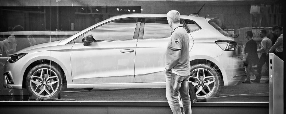 A man holding shopping bags standing outside a vehicle showcase contemplating on whether or not to make a car purchase