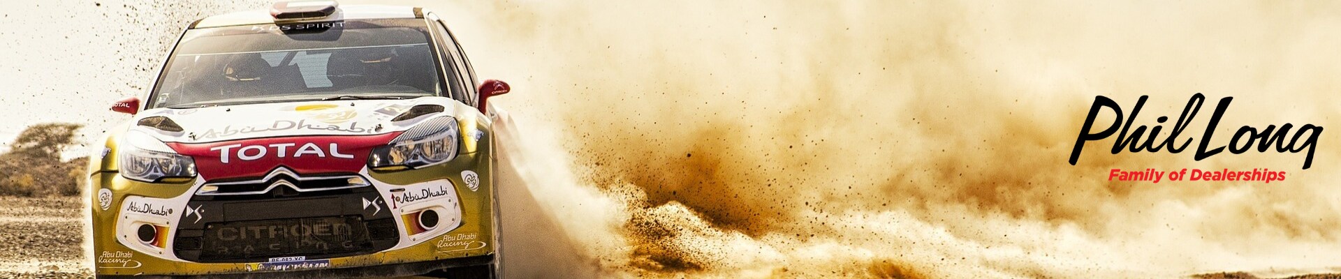 Front view of a rally car drifting through desert sand kicking up a large dust cloud with Phil Long Family of Dealerships logo in the dirt cloud