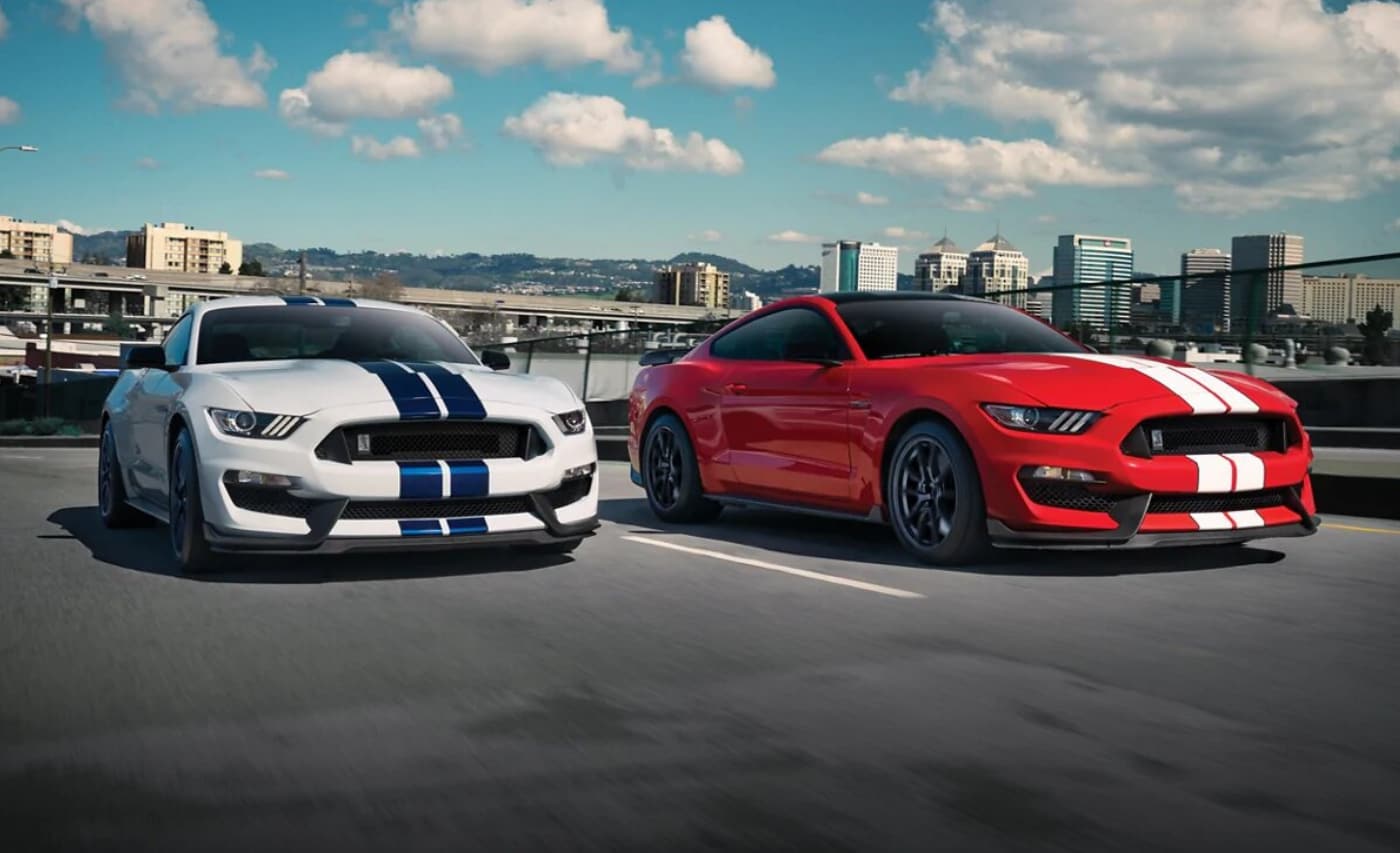 White Ford Mustang Shelby GT350 with blue stripes racing beside a red Ford Mustang Shelby GT350R with white racing stripes