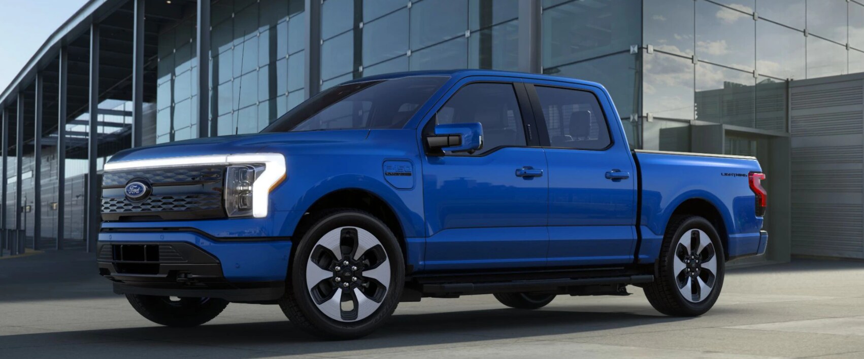 Profile view of a cobalt blue Ford F-150 Lightning with chrome alloy wheels with black cutout detailing and a modern glass and steel building in the background 