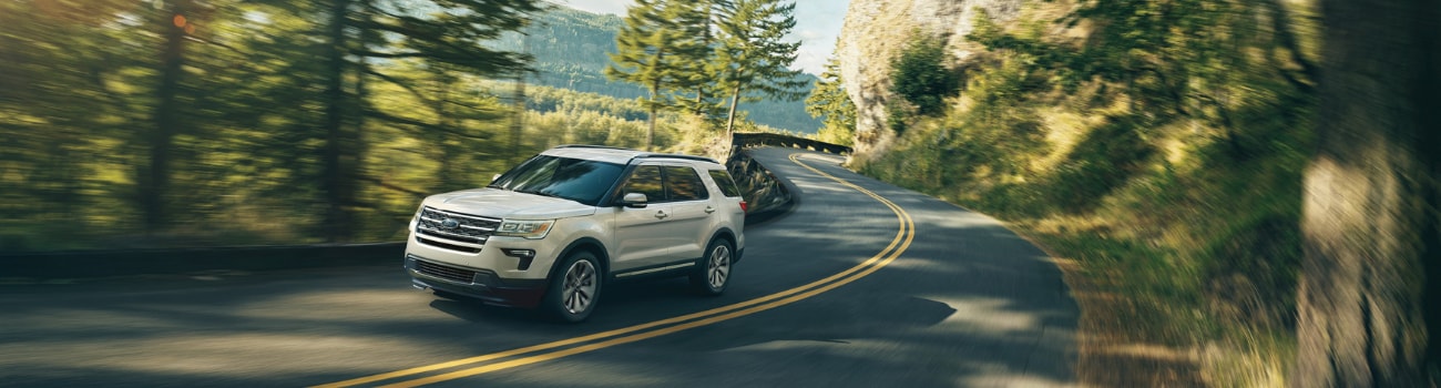 A white 2019 Ford Explorer driving down a curvy road around the edge of a mountain side through a green forest