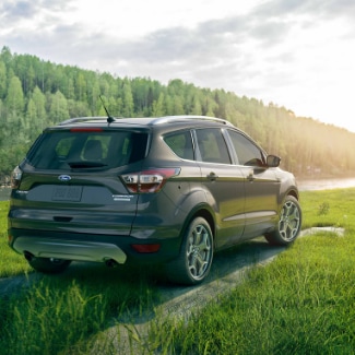A
look at the back side view of a steel green 2019 Ford Escape parked on a grassy
knoll overlooking a river that runs by the entrance of thick green forest