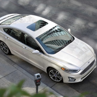 An
aerial view of a white 2019 Ford Fusion parked on the side of a street with a
parking meter and blurred tree leaves in foreground