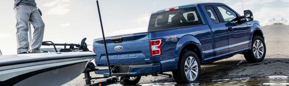Rear angle of a velocity blue 2019 Ford F-150 pulling a fishing boat out of the water with a young man standing in the boat with a fishing pole
