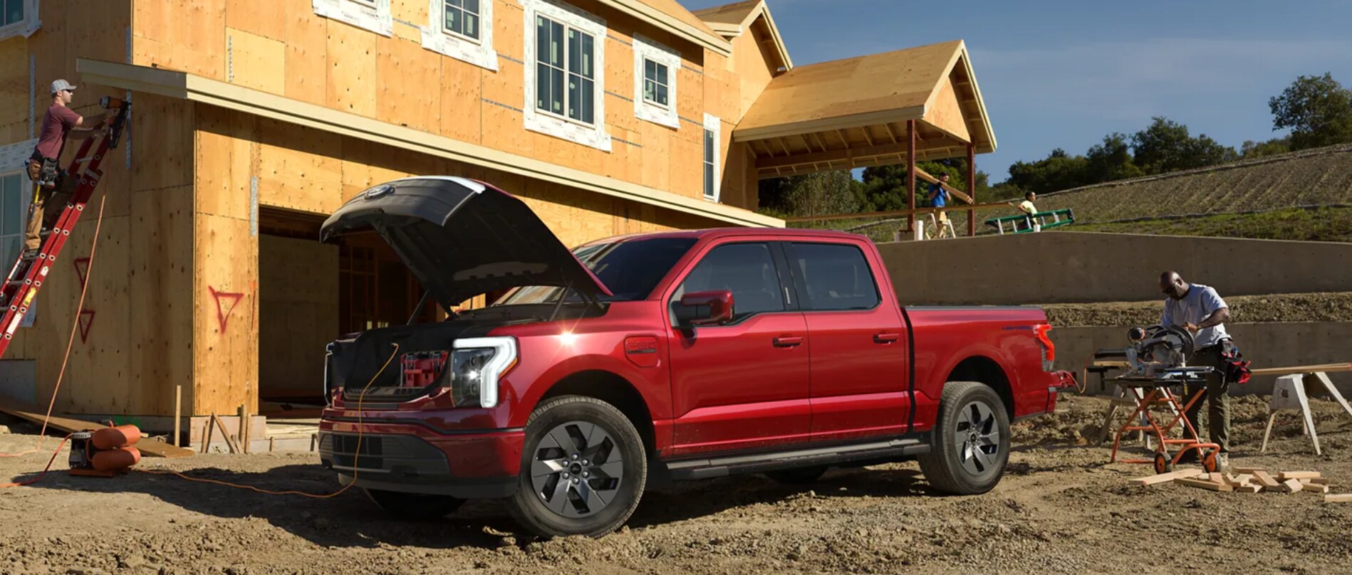 A berry colored F-150 Lightning is parked at a construction site in the
dirt with the hood open revealing a storage space and outlets being used by the
construction workers