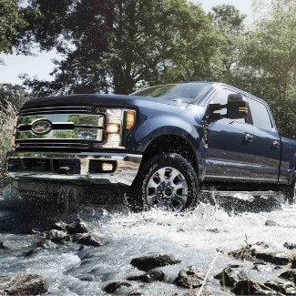 A
front side view of a dark steel blue 2019 Ford Super Duty driving through a
rocky creek with water splashing onto the grill and undercarriage with lush
green trees in the background