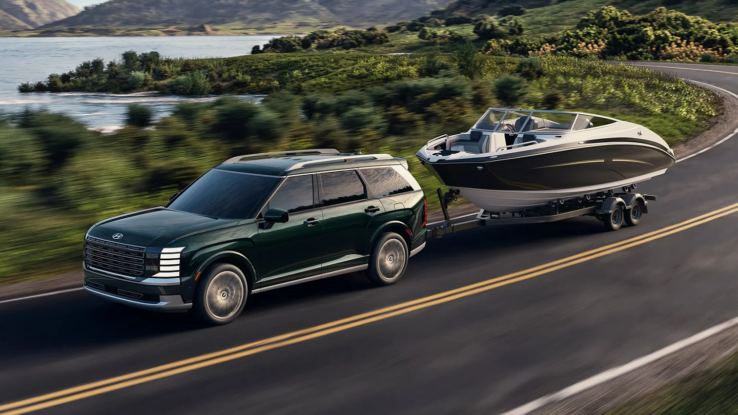2027 Hyundai Palisade in Robust Emerald towing a boat trailer on a winding mountain highway demonstrating 5,000-lb towing capacity