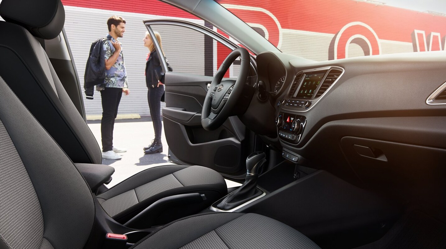 A view through the passenger's door of a 2022 Hyundai Accent through the cabin to the open driver's door reveals a couple standing close by having a conversation and an all dark gray interior and dash