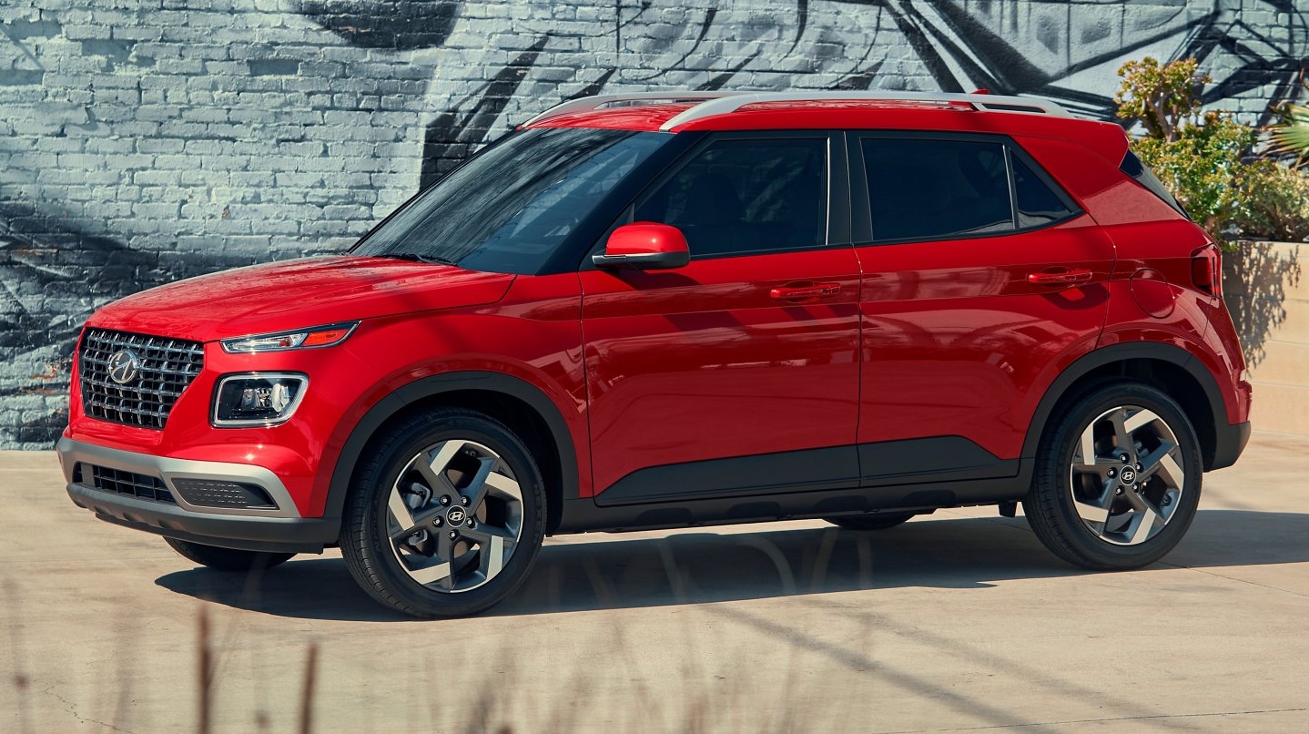 A bright red 2022 Hyundai Venue with dark tinted windows is parked in profile with an angled view of the front of the car during the day in front of an urban graffiti wall