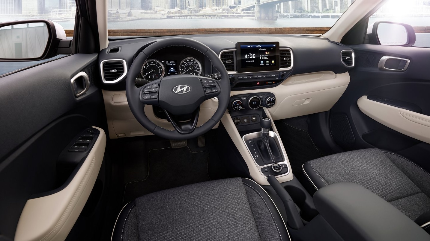 An interior view of the cabin and dash of a 2022 Hyundai Venue with charcoal cloth upholstered seats and a black interior with cream accent panels on the dash and doors, an infotainment screen in the middle of the dash.