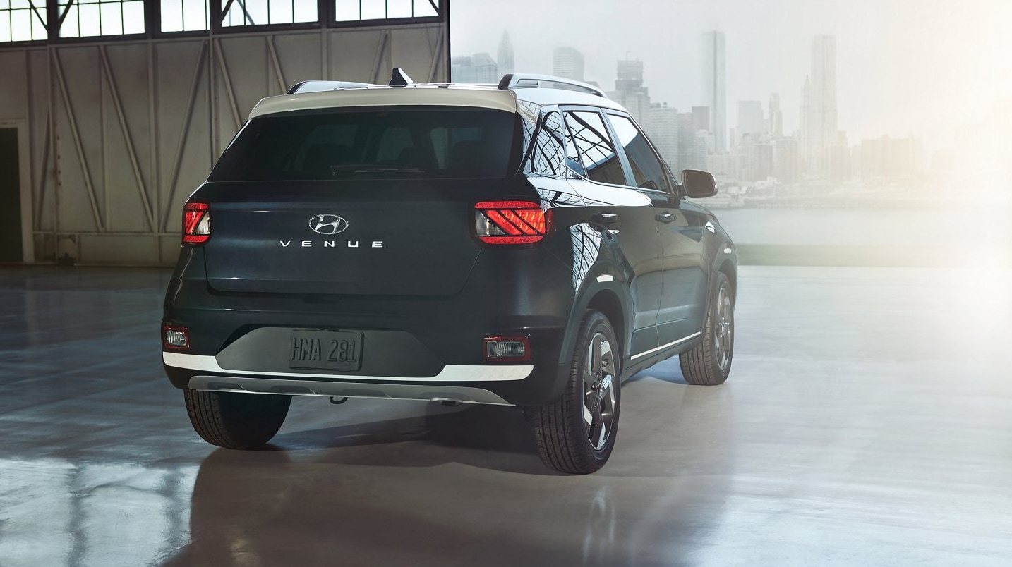 A view of the rear of a dark gray-blue 2022 Hyundai Venue with white rooftop parked in an airplane hangar open to a cityscape on a sunny day.