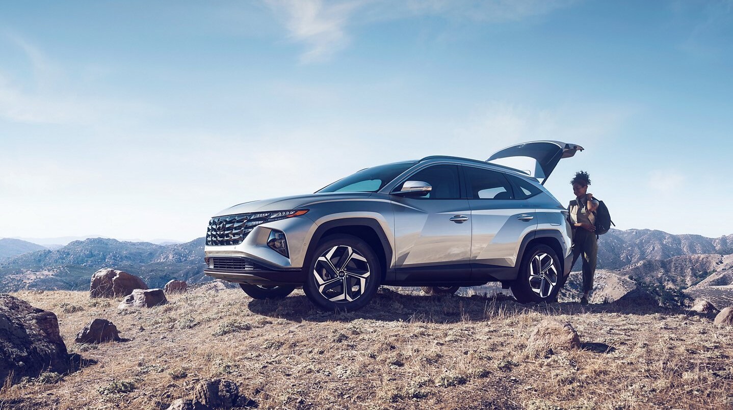 A metallic silver 2022 Hyundai Tucson sits parked on a rocky hill as a woman grabs gear from the open back hatch on a sunny day.