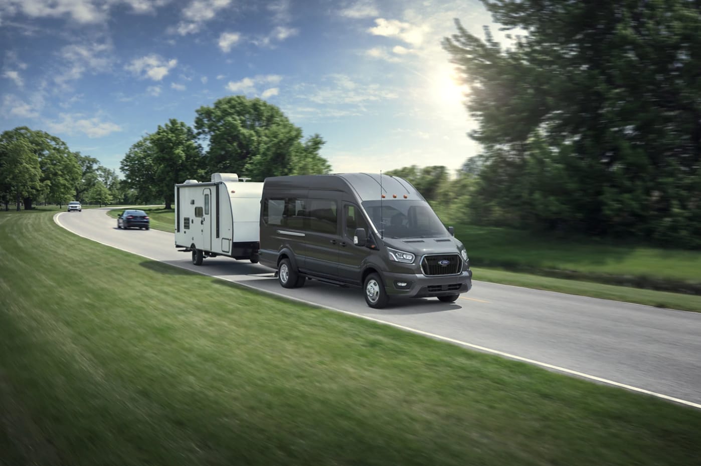 A gray 2023 Ford E-Series Van hauling a camper driving down a country highway