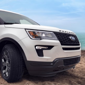 The front passenger side of a white 2019 Ford Explorer parked with the
wheels turned on a sandy beach and crystal clear ocean water in the background