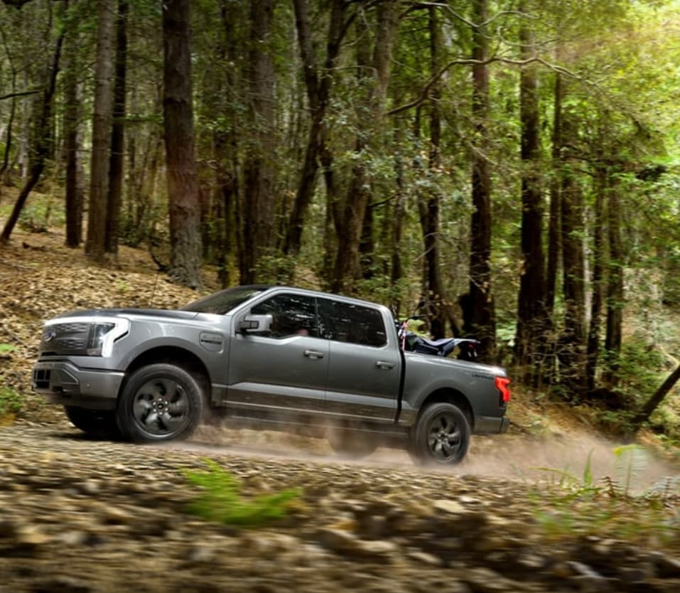 A dark grey Ford F-150 Lightning zooms along a forested road, leaving a cloud of dust in its wake