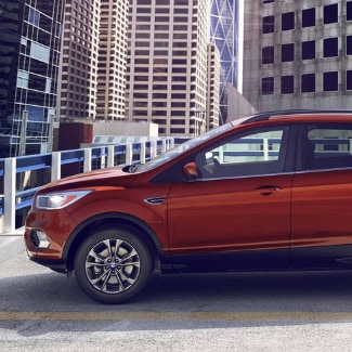 The driver side of an orange 2019 Ford Escape parked on the top floor of
a parking garage in a big city with tall skyscrapers featured in the background