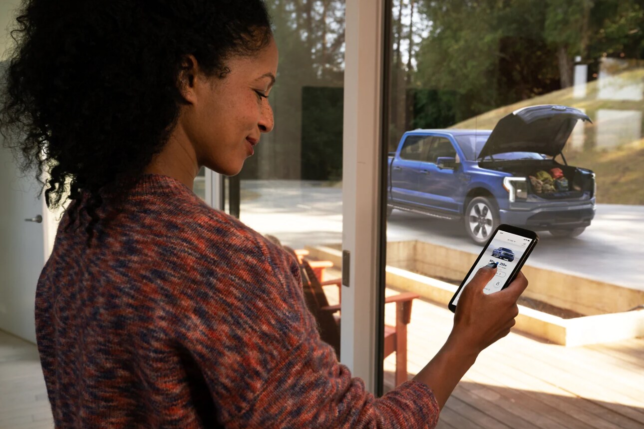 A woman is shown standing in front of a window in her house holding her smartphone which she has just used to open up the front trunk on her Ford F-150 Lightning that is shown just outside