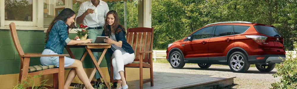 Three friends sitting at a round wooden table outside a country cafe enjoying their meals as a brand new red 2019 Ford Escape awaits them in the parking spot