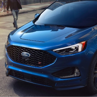 The top driver side view of the front of a blue 2019 Ford Edge pulling
up to a stop sign at a busy city intersection with a pedestrian crossing the
street in the background