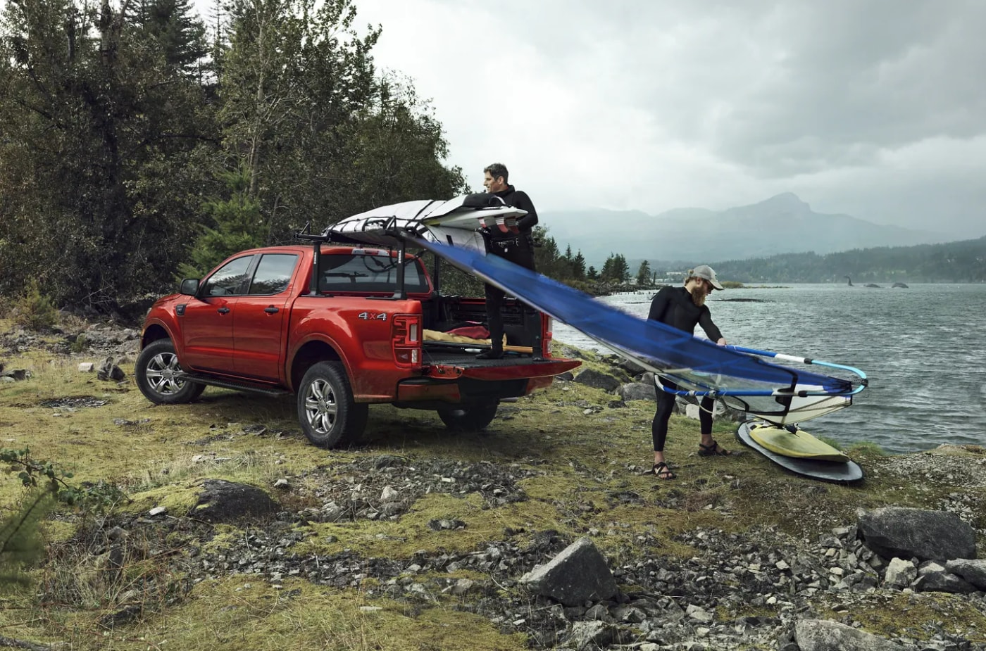 Two men unload windsurfing gear from a red 2023 Ford Ranger parked near a lake