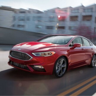 A red 2019 Ford Fusion driving fast down a city highway with the sun
glaring off the hood and a view of blurred apartment buildings in the
background
