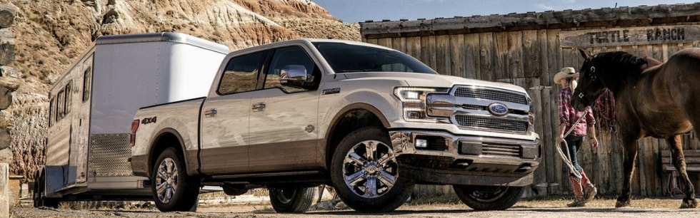Front passenger side angle of an off white 2019 Ford F-150 pulling a horse trailer parked outside of a barn with a woman walking a horse by the Ford truck