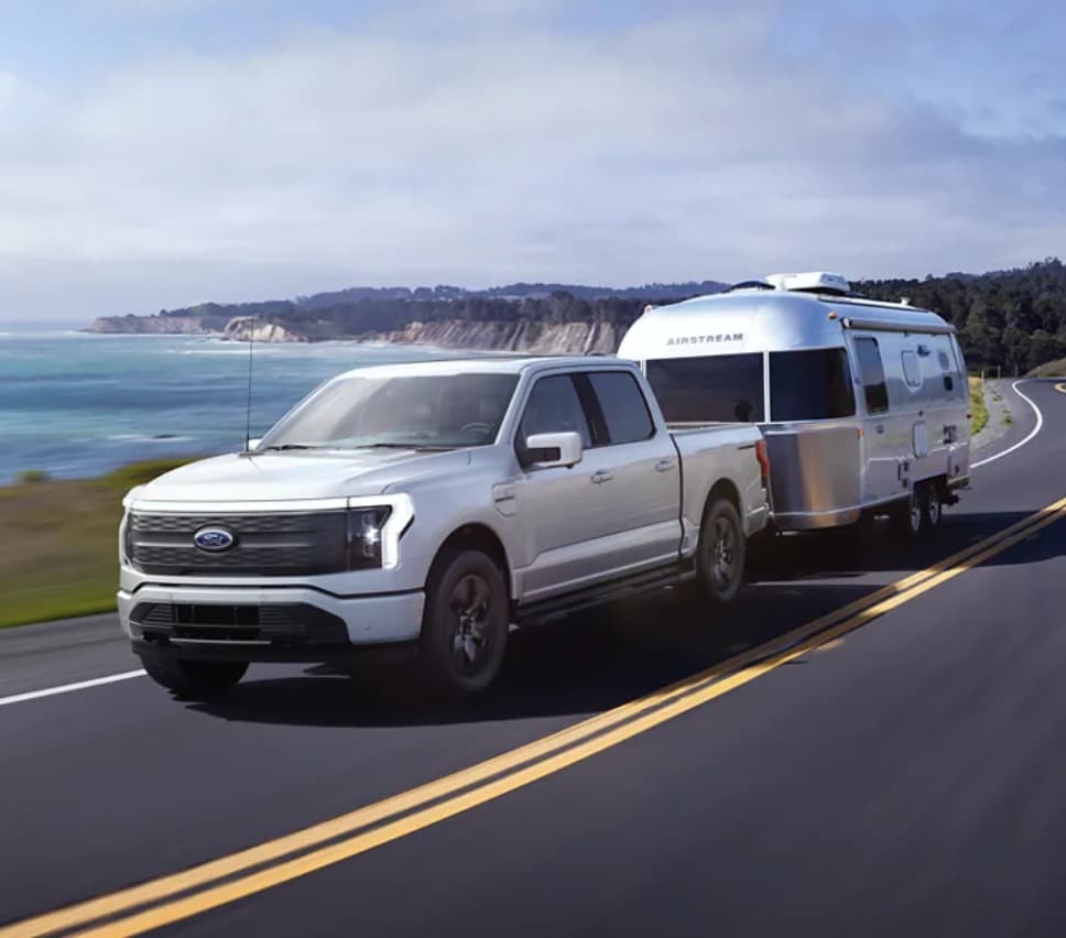 A shiny silver F150 Lightning cruises along a curving coastal 
highway with a silver airstream in tow