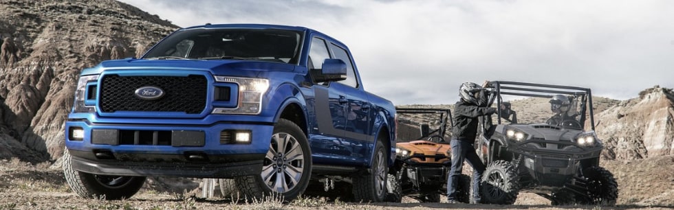 Front angle of a cobalt blue 2019 Ford F-150 headlights shining parked on a desert hill offloading several ATVs