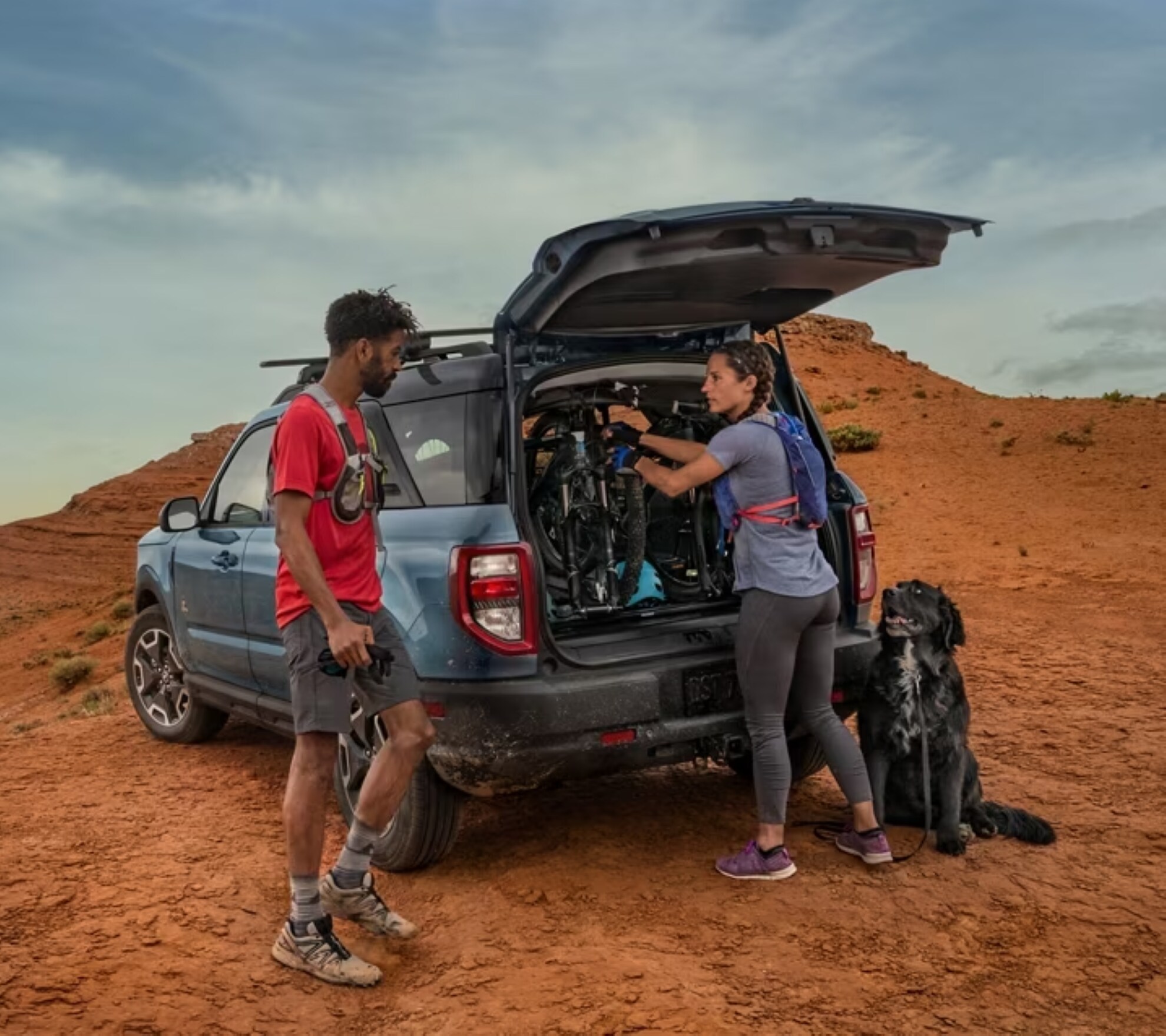 A Ford Bronco parked in a desert with two people setting up a campsite. 