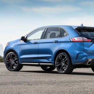 The drivers side view of a blue 2019 Ford Edge parked on a cement
opening surrounded by green bushes with a hazy view of a city far in the distant
background