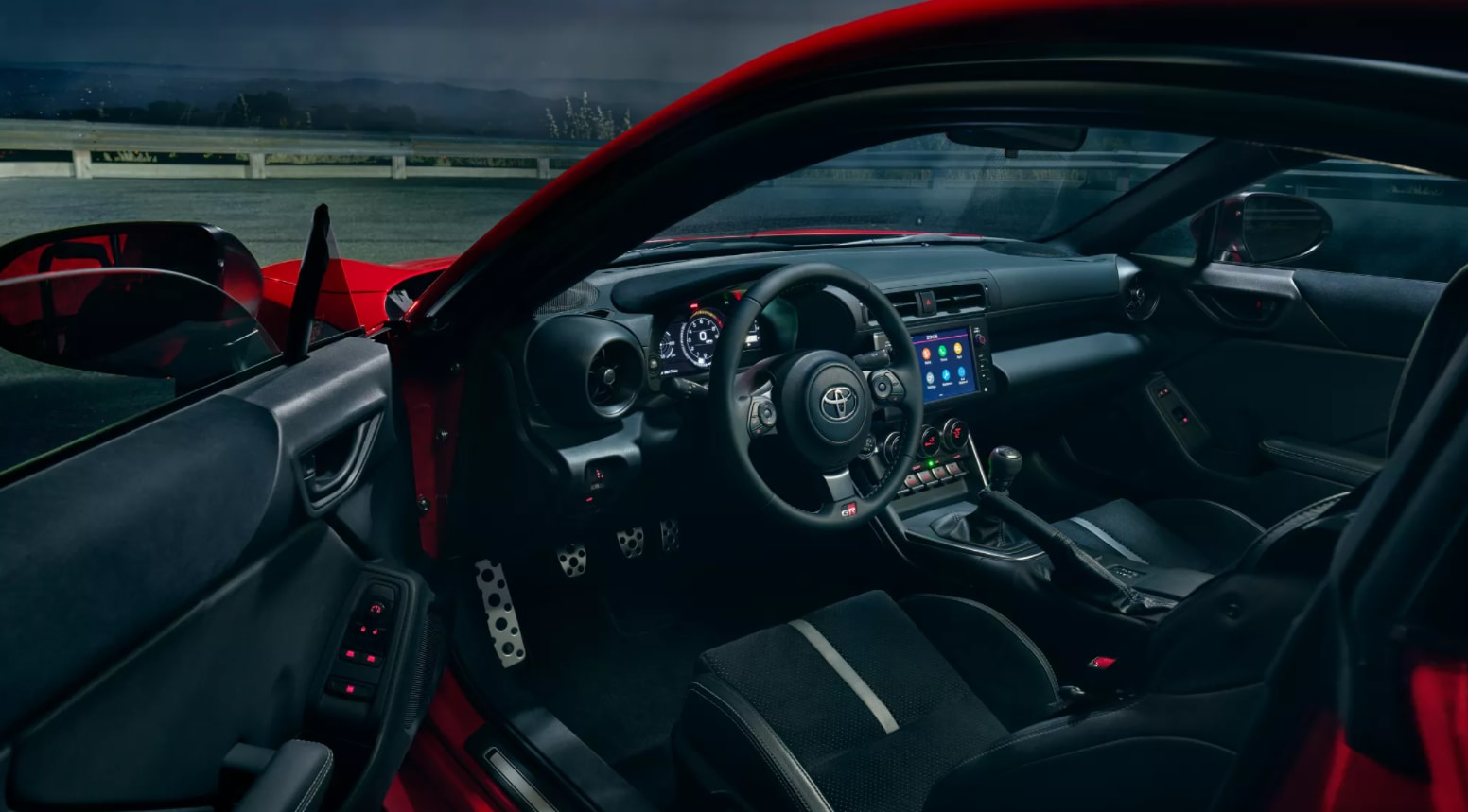 A view into the driver's seat and dash through the open driver's side door shows an all black interior and metal-clad sports pedals