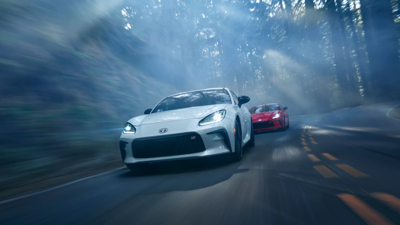 A sleek white Toyota GR86 speeds towards the camera and is passing another car on a two-lane highway through a shadowy forest